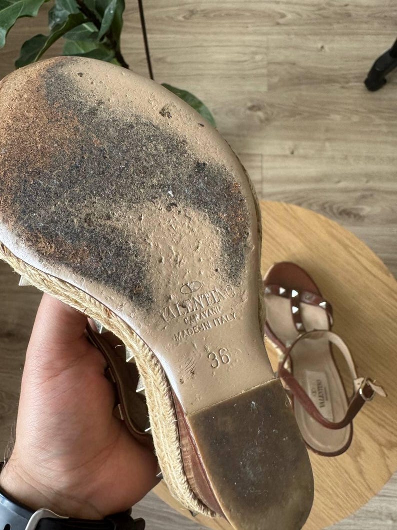 May include: A tan wedge sandal with a jute-wrapped sole and metal studs. The sole is worn and features the embossed text Valentino Garavani Made in Italy and the size 36. A second matching sandal is visible in the background with a brown strap.