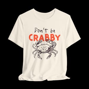 May include: A cream-colored short-sleeve t-shirt featuring a black line-art illustration of a crab centered on the chest. Above the crab, the text reads "Don't be" in black script and "CRABBY" in bold, red, uppercase block letters.
