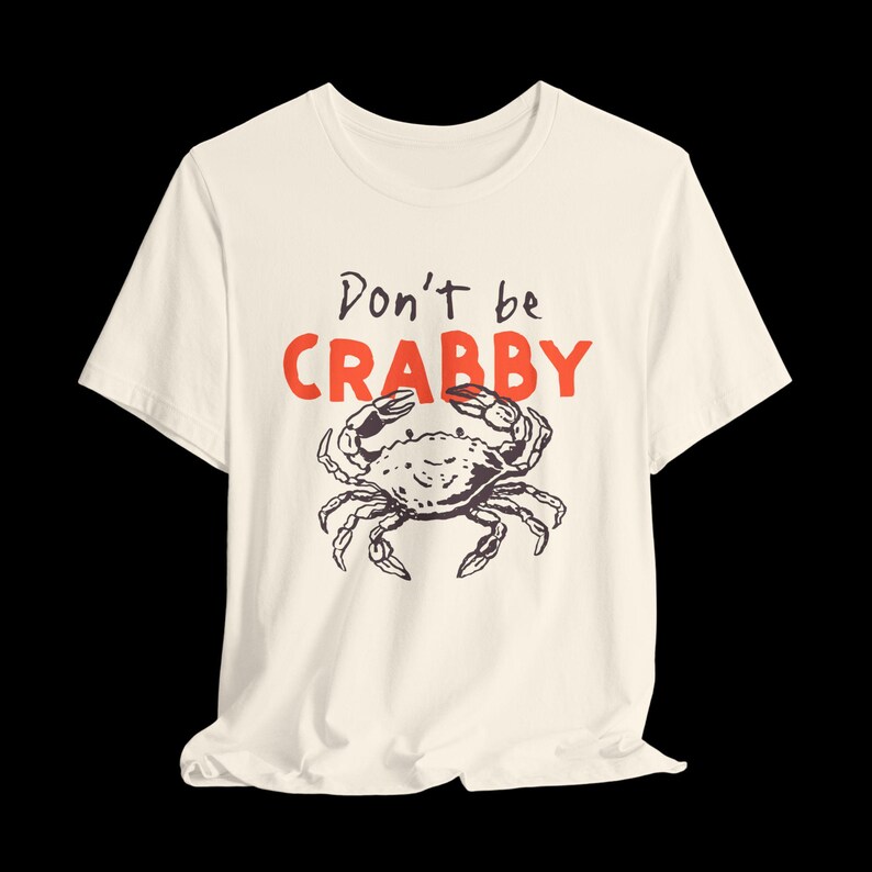 May include: A cream-colored short-sleeve t-shirt featuring a black line-art illustration of a crab centered on the chest. Above the crab, the text reads "Don't be" in black script and "CRABBY" in bold, red, uppercase block letters.