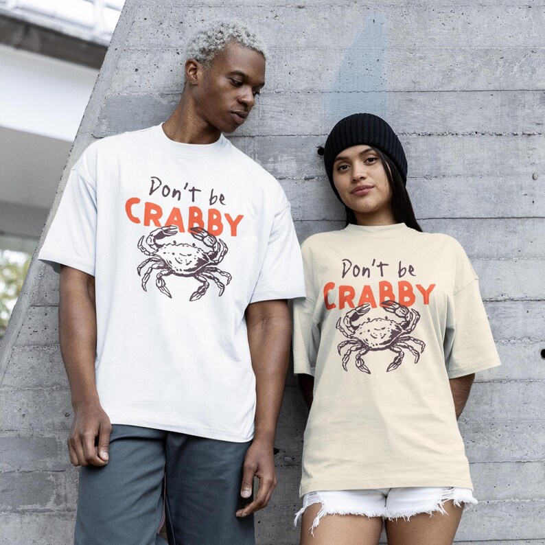 May include: Two short-sleeve t-shirts, one white and one cream, each featuring a black line-art illustration of a crab centered on the chest. Above the crab, the text reads "Don't be" in black script and "CRABBY" in bold, orange capital letters.