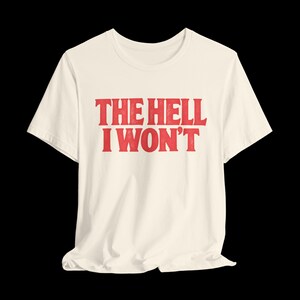 May include: A cream-colored short-sleeve t-shirt featuring the red text THE HELL I WON'T printed in a bold, distressed serif font across the center of the chest. The garment has a classic crew neckline and a standard, relaxed fit silhouette.