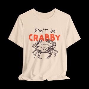 May include: A cream-colored short-sleeve t-shirt featuring a black line-art illustration of a crab centered on the chest. Above the crab, the text reads Don't be in black script and CRABBY in large, bold, orange capital letters.
