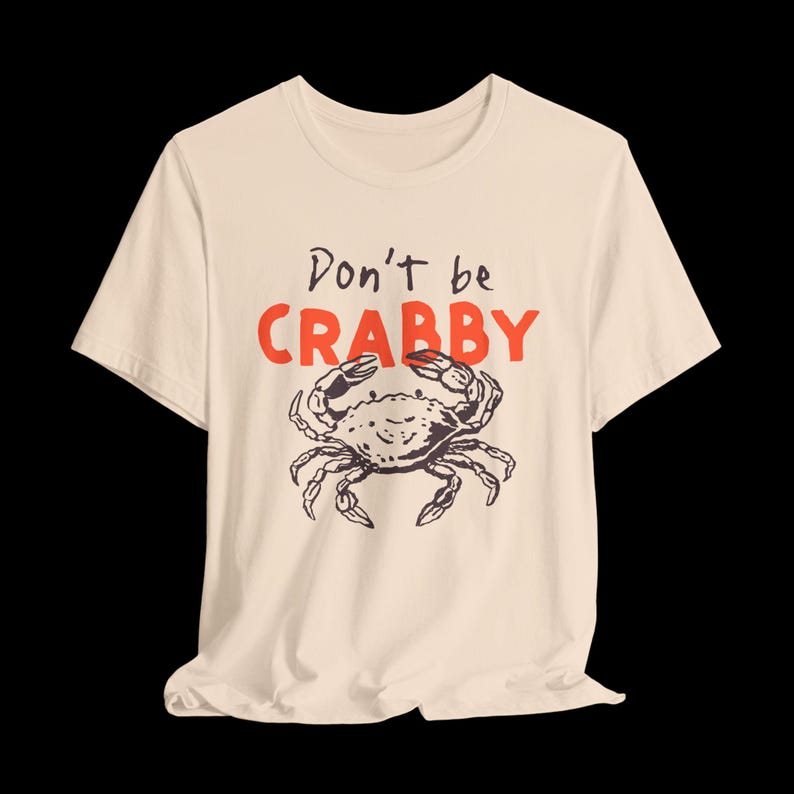 May include: A cream-colored short-sleeve t-shirt featuring a black line-art illustration of a crab centered on the chest. Above the crab, the text reads Don't be in black script and CRABBY in large, bold, orange capital letters.