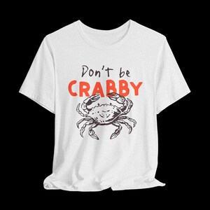 May include: A white short-sleeve t-shirt featuring a black line-art illustration of a crab centered on the chest. Above the crab, the text reads "Don't be" in black script and "CRABBY" in bold, bright orange capital letters.