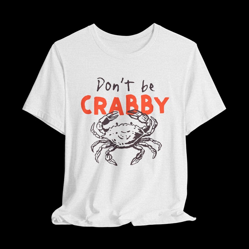 May include: A white short-sleeve t-shirt featuring a black line-art illustration of a crab centered on the chest. Above the crab, the text reads "Don't be" in black script and "CRABBY" in bold, bright orange capital letters.