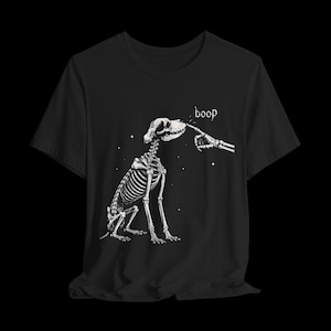 以下が含まれることがあります： 黒いTシャツで、座っている犬の骨格の白いグラフィックイラストが描かれています。骸骨の指が犬の鼻に触れようとしており、指の上にはboopという文字がプリントされています。背景には小さな白い星が散りばめられています。