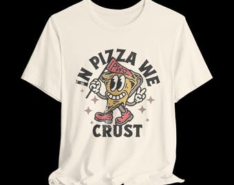 May include: A cream-colored short-sleeve t-shirt featuring a graphic of a cartoon pizza slice character with arms and legs. The text reads IN PIZZA WE CRUST with a small pennant flag labeled IPWC. The design uses black, red, and tan colors.