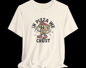 Könnte beinhalten: Cremefarbenes T-Shirt mit einem Cartoon-Pizzastück, das ein lächelndes Gesicht hat, eine Flagge mit der Aufschrift "PWG" hält und ein Friedenszeichen macht. Der Text "IN PIZZA WE CRUST" ist um das Pizzastück herum gedruckt.