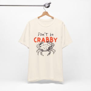 May include: A cream-colored short-sleeve t-shirt featuring a black line-art illustration of a crab centered on the chest. Above the crab, the text reads "Don't be" in black script and "CRABBY" in bold, bright orange capital letters.