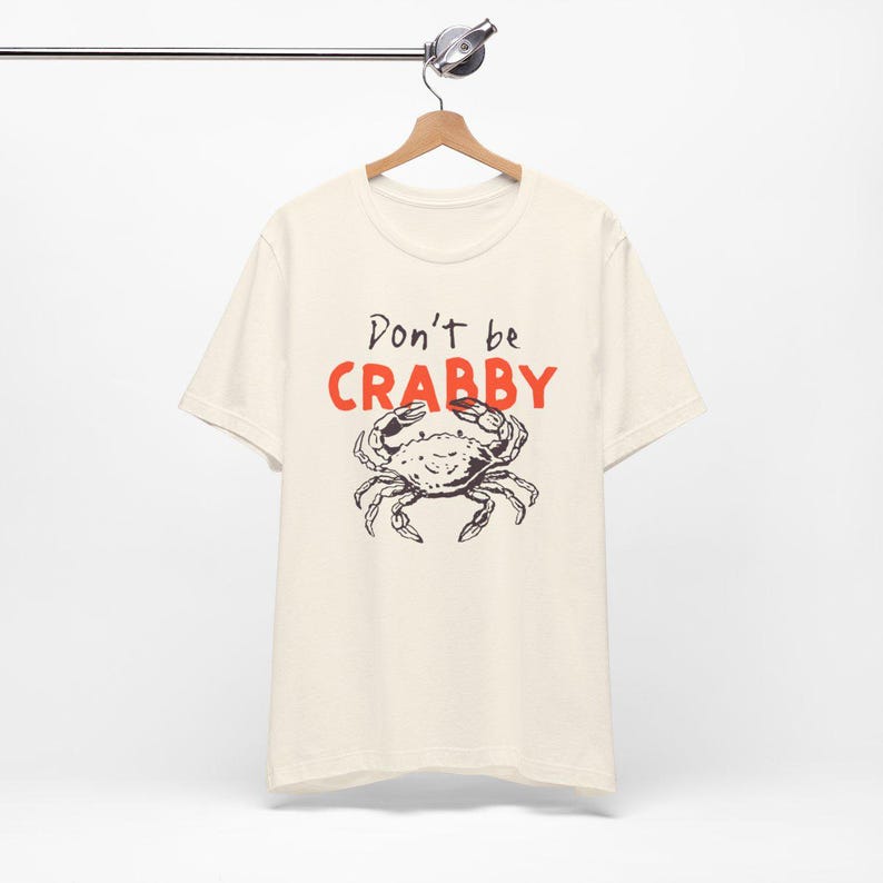 May include: A cream-colored short-sleeve t-shirt featuring a black line-art illustration of a crab centered on the chest. Above the crab, the text reads "Don't be" in black script and "CRABBY" in bold, bright orange capital letters.