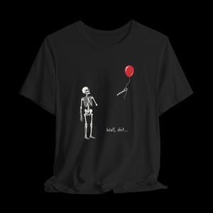 May include: A black short-sleeved t-shirt featuring a graphic of a white skeleton looking at a detached skeletal hand holding a red balloon. The text "Well, shit..." is printed in white below the skeleton. The design is centred on the chest.