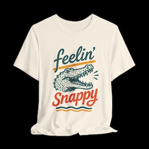 May include: A cream-coloured short-sleeve t-shirt featuring a graphic of an alligator head with its mouth open. The text "feelin'" appears in dark teal above the alligator, and "Snappy" is written in red script below, accented by orange lines.