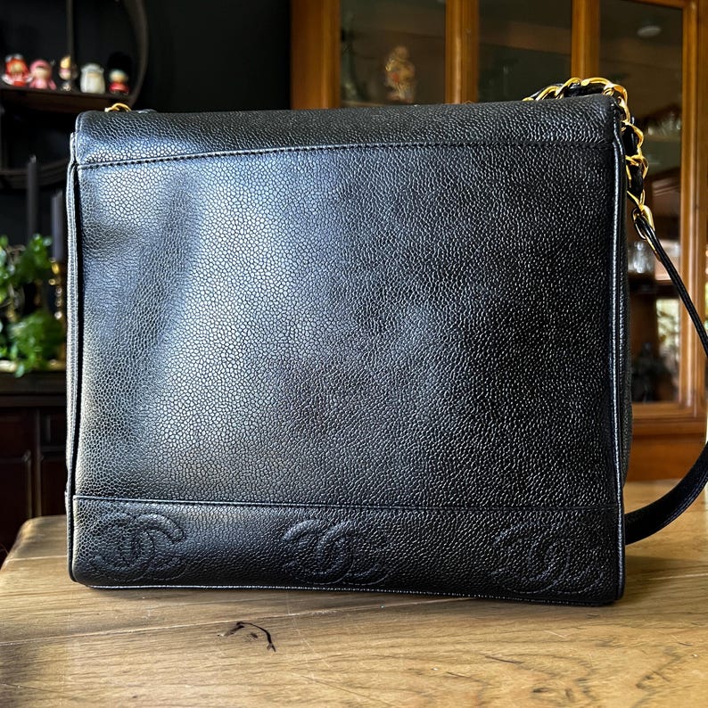 May include: Black leather shoulder bag with a textured surface and gold-tone chain strap. The bag features embossed interlocking Cs along the bottom edge. The bag is sitting on a wooden surface.