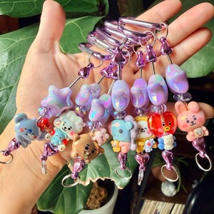 May include: A collection of six beaded lanyards featuring metallic purple carabiner clips. Each lanyard is decorated with iridescent purple star and oval beads, small clear spacers, and a unique cartoon character charm in various colors.