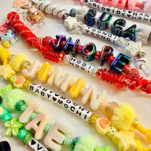 May include: A collection of colorful beaded lanyards featuring large letter beads spelling names like JIN, SUGA, JHOPE, JIMIN, and TAE. The lanyards include various charms, fuzzy pom-poms, star-shaped beads, and smaller letter beads with phrases like WORLDWIDE HANDSOME and BABY MOCHI.