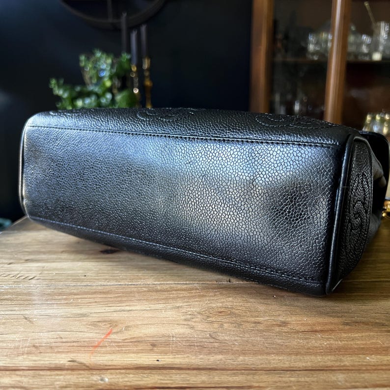 May include: Black leather handbag with a textured surface. The bag features embossed detailing and a structured shape. The bottom of the bag is visible, resting on a wooden surface.