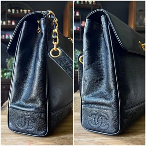 May include: Black leather handbag with gold-tone chain straps and hardware. The bag features a flap closure and embossed logo details on the bottom corners. The leather has a textured appearance.