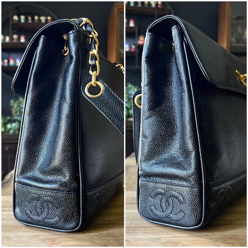 May include: Black leather handbag with gold-tone chain straps and hardware. The bag features a flap closure and embossed logo details on the bottom corners. The leather has a textured appearance.