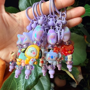Pode incluir: Uma coleção de porta-chaves roxos com contas iridescentes em forma de estrela e lua, contas roxas facetadas transparentes e vários berloques de personagens de desenhos animados, incluindo uma figura com capuz amarelo, um coala azul e um coração vermelho.