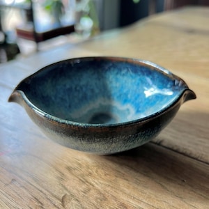 May include: A small, blue ceramic bowl with a textured, dark brown rim. The bowl has a slightly flared lip and a unique, asymmetrical shape.