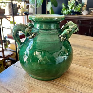 May include: A green ceramic vase with two handles shaped like frogs. The vase has a crackled glaze and a glossy finish.