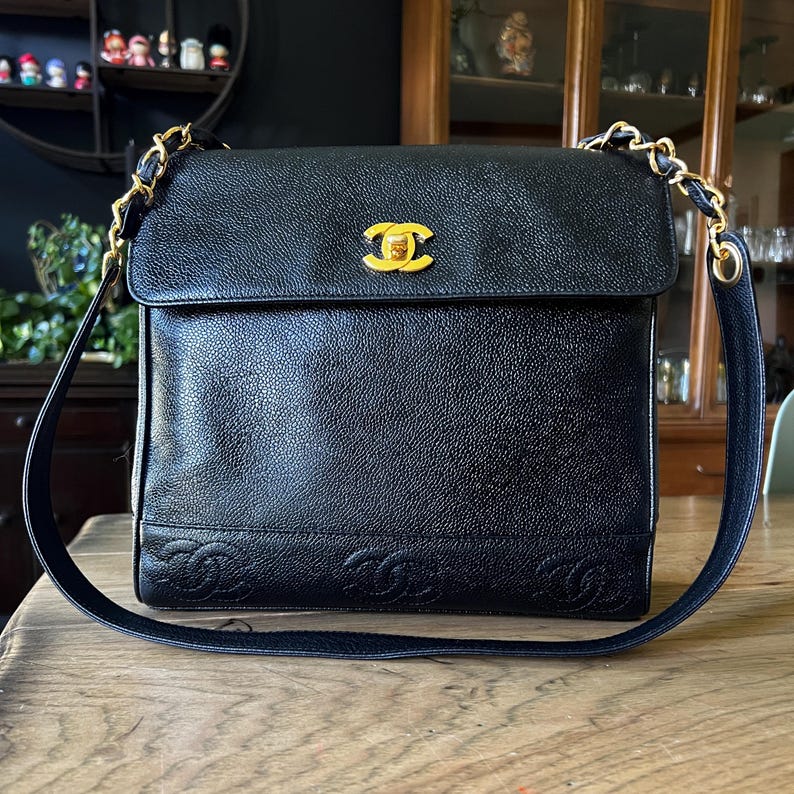 May include: Black leather shoulder bag with a gold-tone chain strap. The bag features a flap closure with a gold logo and embossed logo details at the bottom. The bag is sitting on a wooden surface.