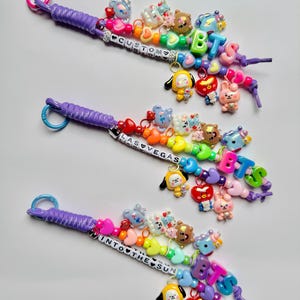 May include: Three beaded lanyards featuring purple paracord, colorful plastic letter beads spelling CUSTOM, LAS VEGAS, and INTO THE SUN, and various character charms including hearts, stars, and small figures in vibrant shades of pink, blue, and yellow.