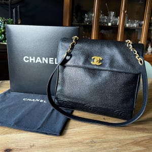 May include: Black leather Chanel shoulder bag with a gold chain strap and a gold logo clasp. The bag has a flap closure and a textured surface. A black box and dust bag with the Chanel logo are also present.