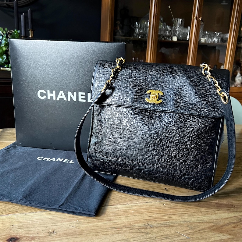 May include: Black leather Chanel shoulder bag with a gold chain strap and a gold logo clasp. The bag has a flap closure and a textured surface. A black box and dust bag with the Chanel logo are also present.