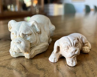 Può includere: Due statuine di bulldog in ceramica bianca. La statuina più grande è sdraiata con la testa appoggiata sulle zampe. La statuina più piccola è sdraiata con la testa girata di lato. Entrambe le statuine hanno dettagli realistici.
