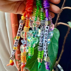 May include: Three colorful beaded lanyards made of braided cord in red, green, and purple. The beads include clear, iridescent, and striped shapes. One lanyard features the text INTO THE SUN, while another displays BORAHAE ALIENS with heart icons.