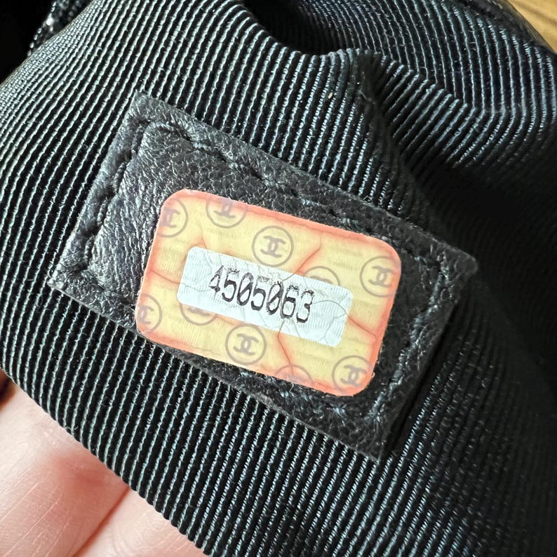 May include: Close-up of a black fabric with a stitched pattern. A rectangular label with the number "4505063" is visible. The label has a yellow background with the Chanel logo.