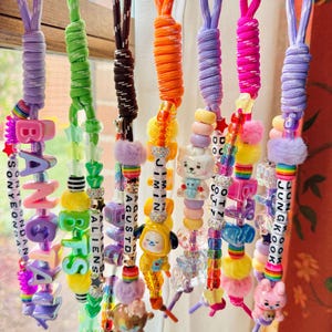 May include: Eight colorful lanyards hanging from a white rod, each featuring paracord loops, plastic letter beads spelling names like BANGTAN, JIMIN, and JUNGKOOK, along with various charms, fuzzy pom-poms, and vibrant plastic beads in many shapes.