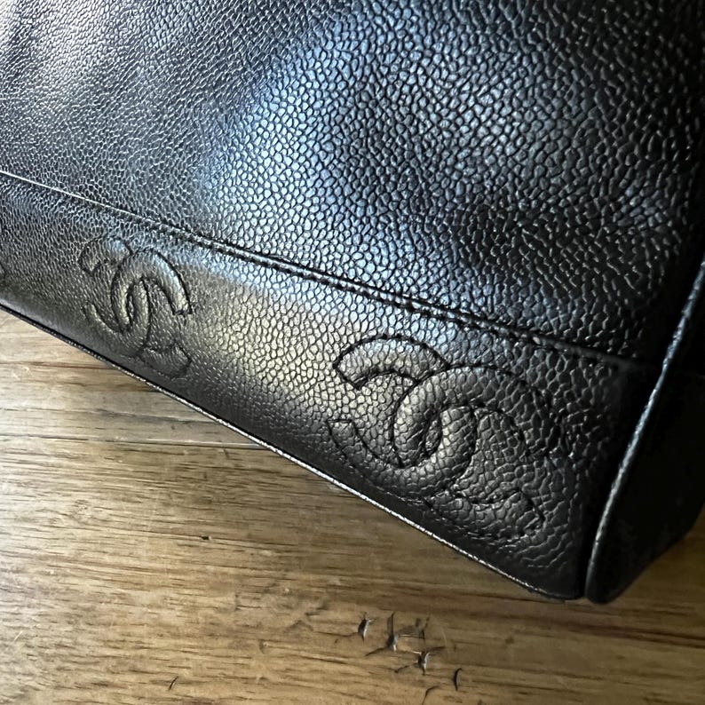May include: Close-up of a black leather handbag with a textured surface. The bag features embossed interlocking double C logos along the bottom edge, a signature design element.