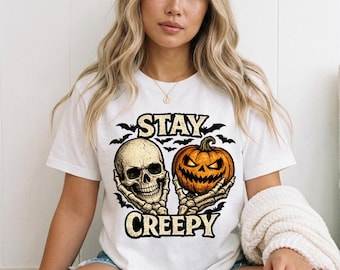 May include: A white short-sleeve t-shirt featuring a graphic print of two skeletal hands holding a human skull and a carved orange jack-o'-lantern. The text STAY CREEPY is printed in a bold, distressed font above and below the central illustration.