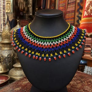 Pode incluir: Um colar de contas multicolorido com um padrão vibrante em ziguezague. O colar é feito com pequenas contas redondas nas cores amarela, preta, branca, verde, azul e laranja, dispostas em fileiras geométricas repetitivas que formam uma peça decorativa larga.