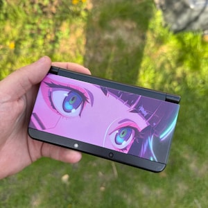 May include: A black handheld gaming console with a pink and purple anime-style graphic of large eyes. The console is held against a background of green grass.