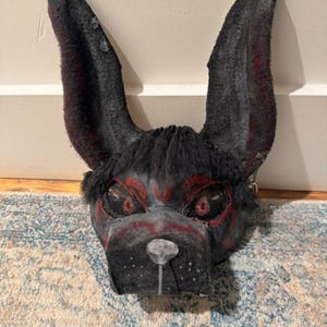 May include: A black and red dog mask with large, upright ears. The mask features red accents around the eyes and on the snout, with a textured, fur-like appearance. The mask is designed to be worn.