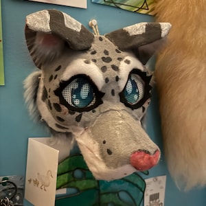 May include: A handcrafted animal mask featuring grey and white fur with dark grey spots. The mask has large, expressive blue eyes with mesh detailing, pointed ears with grey tips, and a pink nose. The design includes white fur accents above the eyes.