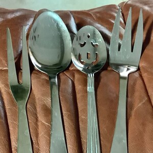 May include: A set of four stainless steel serving utensils. Includes a serving fork, a large spoon, a slotted spoon, and a serving fork. Each utensil has a long handle with a hole at the end. The utensils are on a brown surface.