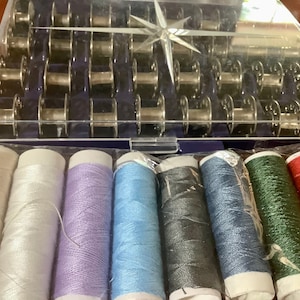 May include: A sewing kit with a clear plastic case containing numerous metal bobbins and a variety of colorful thread spools. The thread colors include white, lavender, blue, black, green, red, and yellow. The kit is designed for sewing and crafting.