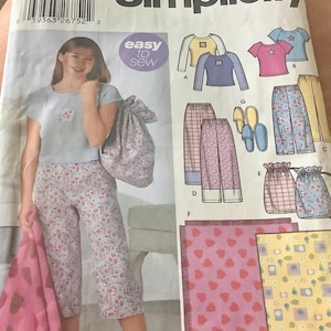 May include: Simplicity sewing pattern 5522, size AA 8-16. The pattern features pajama sets with floral and heart designs. The image shows a woman wearing a pajama set. The pattern includes tops, pants, and accessories. The text "easy to sew" is visible.