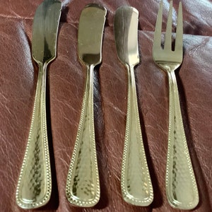 May include: Four pieces of gold-colored flatware. Three butter knives with rounded blades and a fork with three tines. The handles have a textured, hammered design with a beaded border.