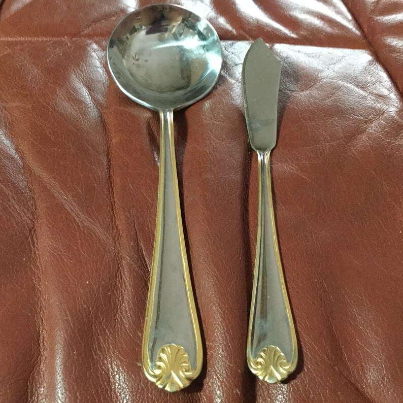 May include: A stainless steel soup spoon and butter knife set. The handles have a gold-colored trim and a decorative gold-colored flourish at the base. The spoon has a round bowl, and the knife has a rounded blade.