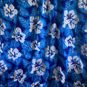 May include: A vibrant blue fabric featuring a repeating pattern of white hibiscus flowers and light blue leaves. The background is a darker shade of blue, creating a tropical floral design.