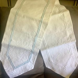 May include: White table runner with a textured surface and light blue stitching along the edges. The runner has a rectangular shape with a decorative design at the ends. This elegant table runner is perfect for adding a touch of sophistication to any dining table.