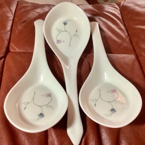 May include: Three white ceramic spoon rests with a floral design. Each rest has a wide, shallow bowl and a long handle. The floral design features delicate purple and blue flowers with green stems and tendrils.