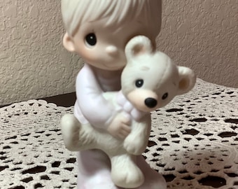 May include: A porcelain figurine of a child in a white outfit holding a white teddy bear. The figurine has a round base and is painted in soft pastel colors. The child's hair is styled in a simple, smooth fashion.