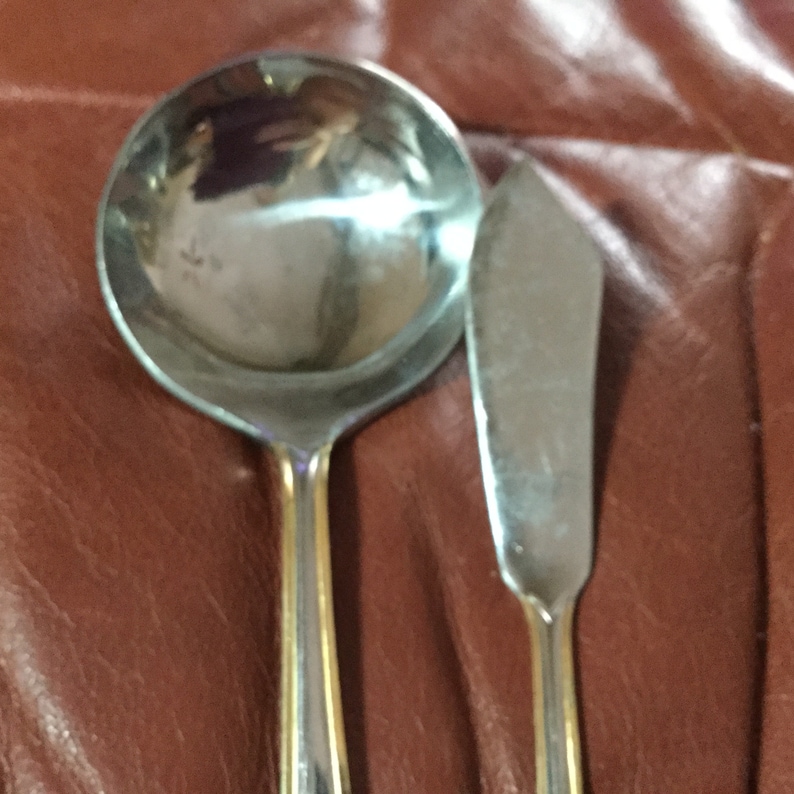 May include: A stainless steel ladle and butter knife set with gold-colored accents on the handles. The ladle has a round bowl, and the butter knife has a flat, angled blade. The set is displayed on a brown surface.