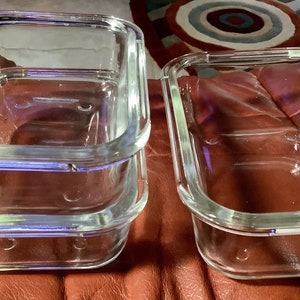 May include: A stack of three clear glass rectangular food storage containers with rounded corners. Another similar container is next to the stack. The containers have a raised rim and small circular indentations on the bottom.